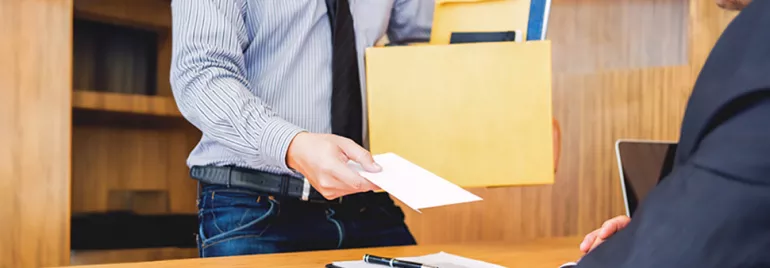 Un empleado, vestido con camisa y corbata, le da una carta de renuncia voluntaria a su jefe mientras sostiene una caja con documentos.