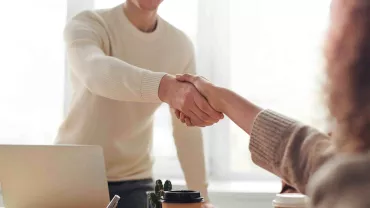 Dos personas se dan la mano sobre una mesa con una laptop y un café.