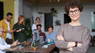 Persona con gafas sonríe en oficina con compañeros al fondo.