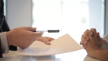 Mano entregando un bolígrafo y documento en un escritorio.