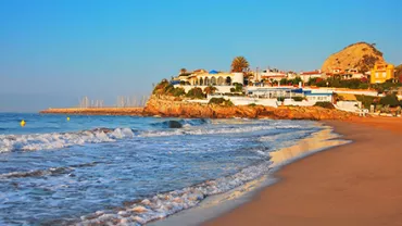 Vista de playa con olas suaves y casas en el fondo al atardecer.