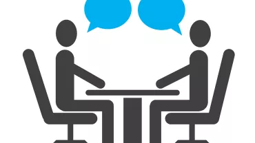 Dos figuras sentadas a una mesa conversando.