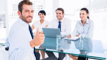 Persona sonriendo con pulgar arriba, grupo de colegas al fondo.