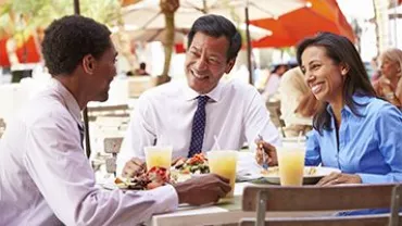 Tres personas sonríen mientras comen en una terraza al aire libre.