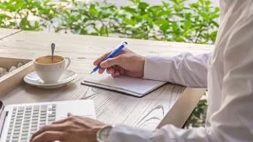 Persona escribiendo en una libreta junto a una taza de café en una mesa de madera.