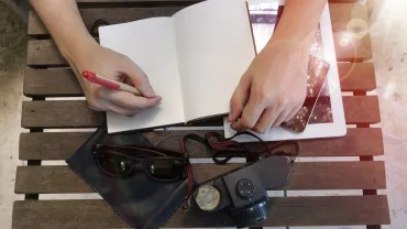 Persona escribiendo en un cuaderno junto a una tableta, cámara y gafas sobre mesa de madera.