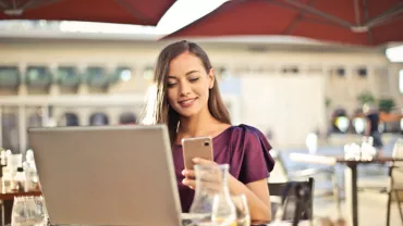 Persona trabajando en laptop y usando teléfono en una terraza.