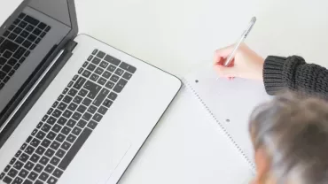 Persona escribiendo en una libreta junto a una computadora portátil.