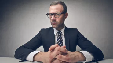 Persona con traje y gafas, manos entrelazadas sobre la mesa.