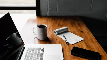 Mesa con portátil, taza de café y cuaderno con bolígrafo.