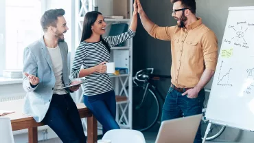 Grupo de personas celebrando en una oficina con una pizarra y una laptop.