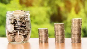 Un frasco lleno de monedas al lado de pilas de monedas en aumento.