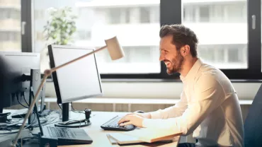 Persona sonriendo mientras trabaja en la computadora en una oficina luminosa.