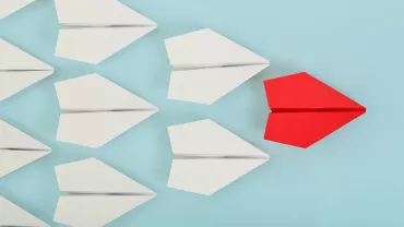 Aviones de papel blancos y uno rojo sobre fondo azul.