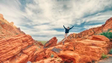 Persona saltando sobre rocas rojizas bajo un cielo parcialmente nublado.