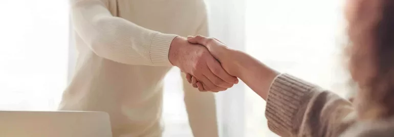 Dos personas se dan la mano sobre una mesa con una laptop y un café.
