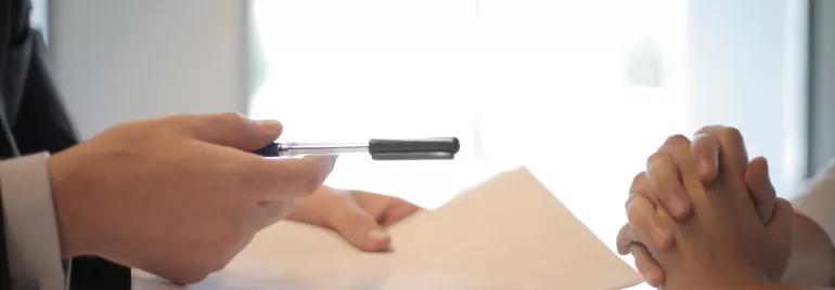 Mano entregando un bolígrafo y documento en un escritorio.
