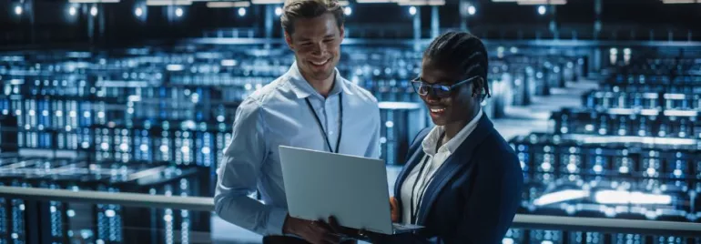 Dos personas revisan una laptop en un centro de datos iluminado.