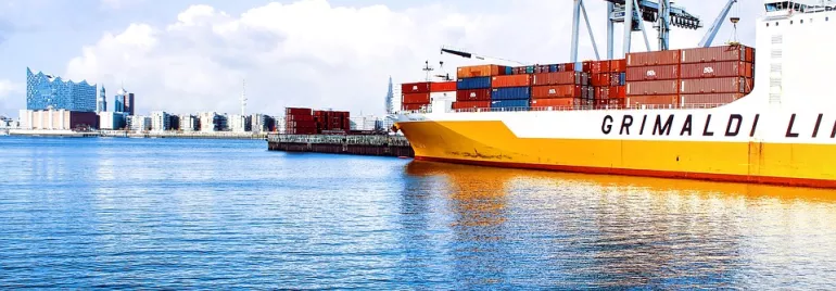 Barco de carga en puerto con contenedores y ciudad al fondo.
