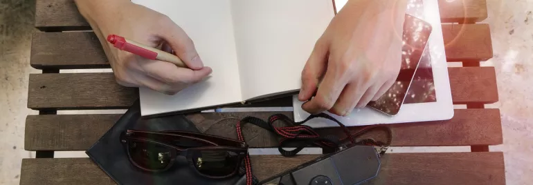 Persona escribiendo en un cuaderno junto a una tableta, cámara y gafas sobre mesa de madera.