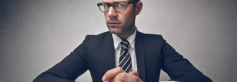Persona con traje y gafas, manos entrelazadas sobre la mesa.