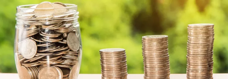 Un frasco lleno de monedas al lado de pilas de monedas en aumento.